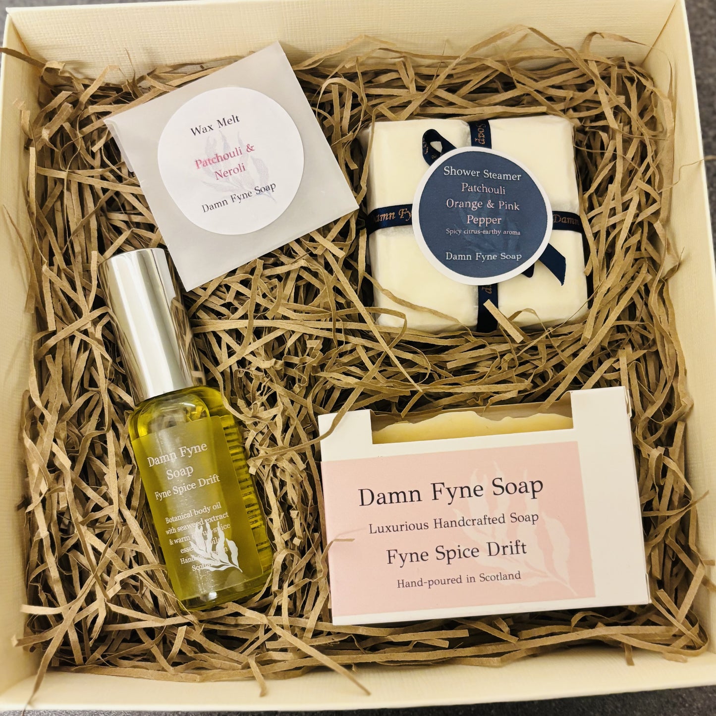 Cream gift box with navy ribbon containing Damn Fyne Soap products: Fyne Spice Drift soap, Fyne Spice Drift body oil, four shower steamers, and a patchouli & neroli wax melt, arranged on shredded paper filler for festive gifting