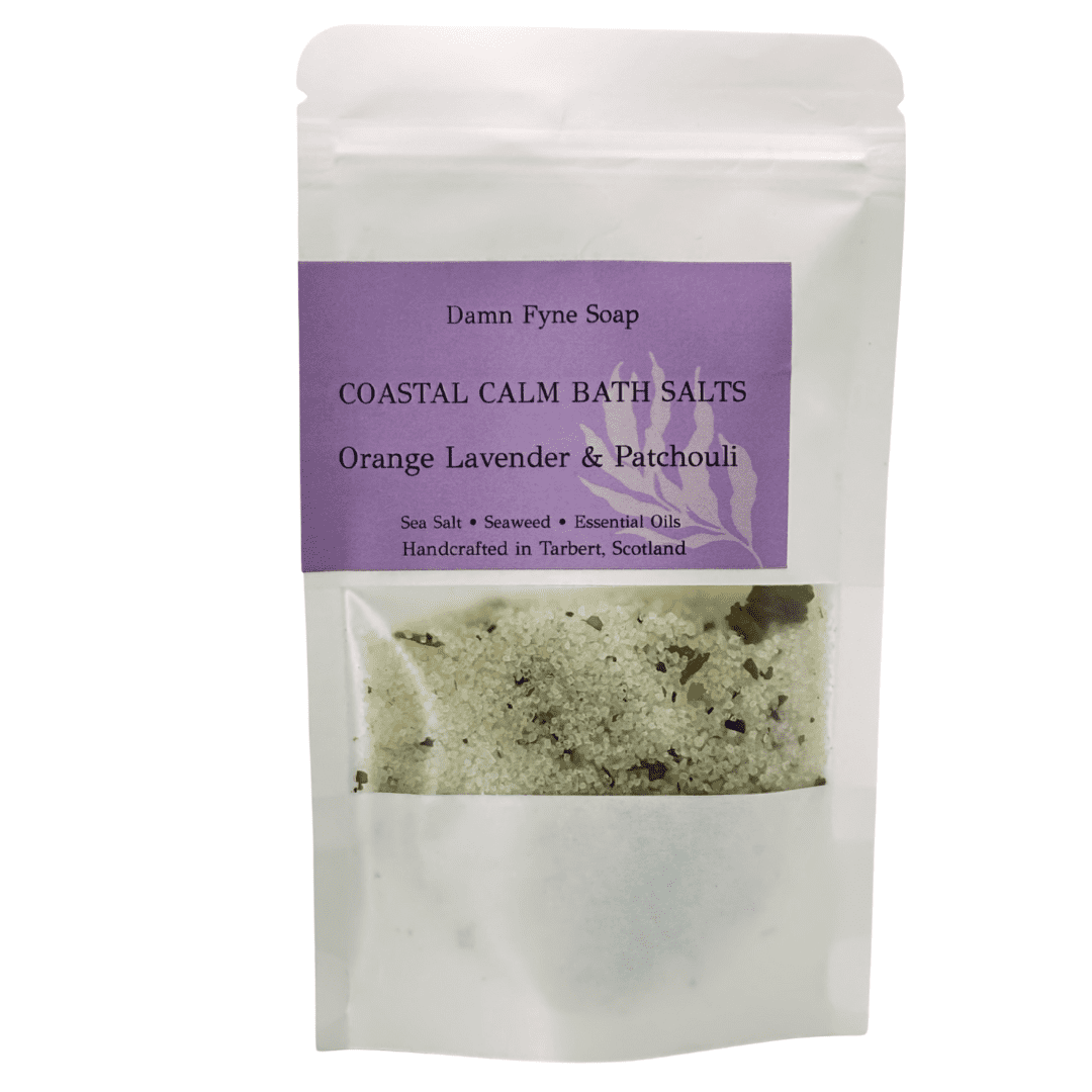 Coastal Calm Bath Salts in a semi-transparent kraft pouch with a purple label. Label reads “Coastal Calm Bath Salts – Orange, Lavender & Patchouli – Sea Salt • Seaweed • Essential Oils – Handcrafted in Tarbert, Scotland.” Contents visible through window, showing coarse salt and botanical elements.