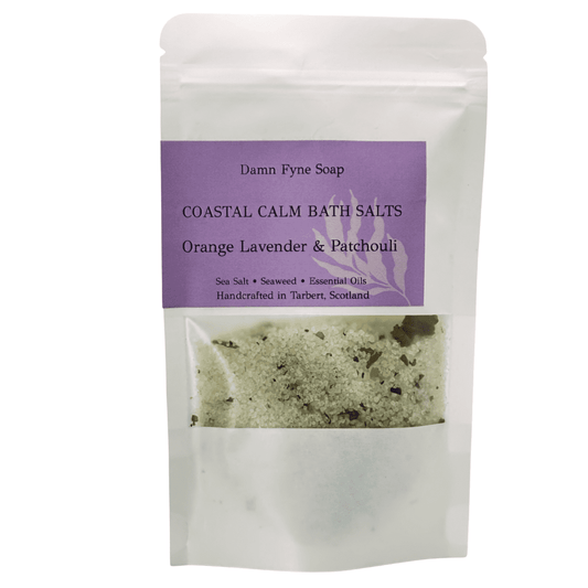 Coastal Calm Bath Salts in a semi-transparent kraft pouch with a purple label. Label reads “Coastal Calm Bath Salts – Orange, Lavender & Patchouli – Sea Salt • Seaweed • Essential Oils – Handcrafted in Tarbert, Scotland.” Contents visible through window, showing coarse salt and botanical elements.