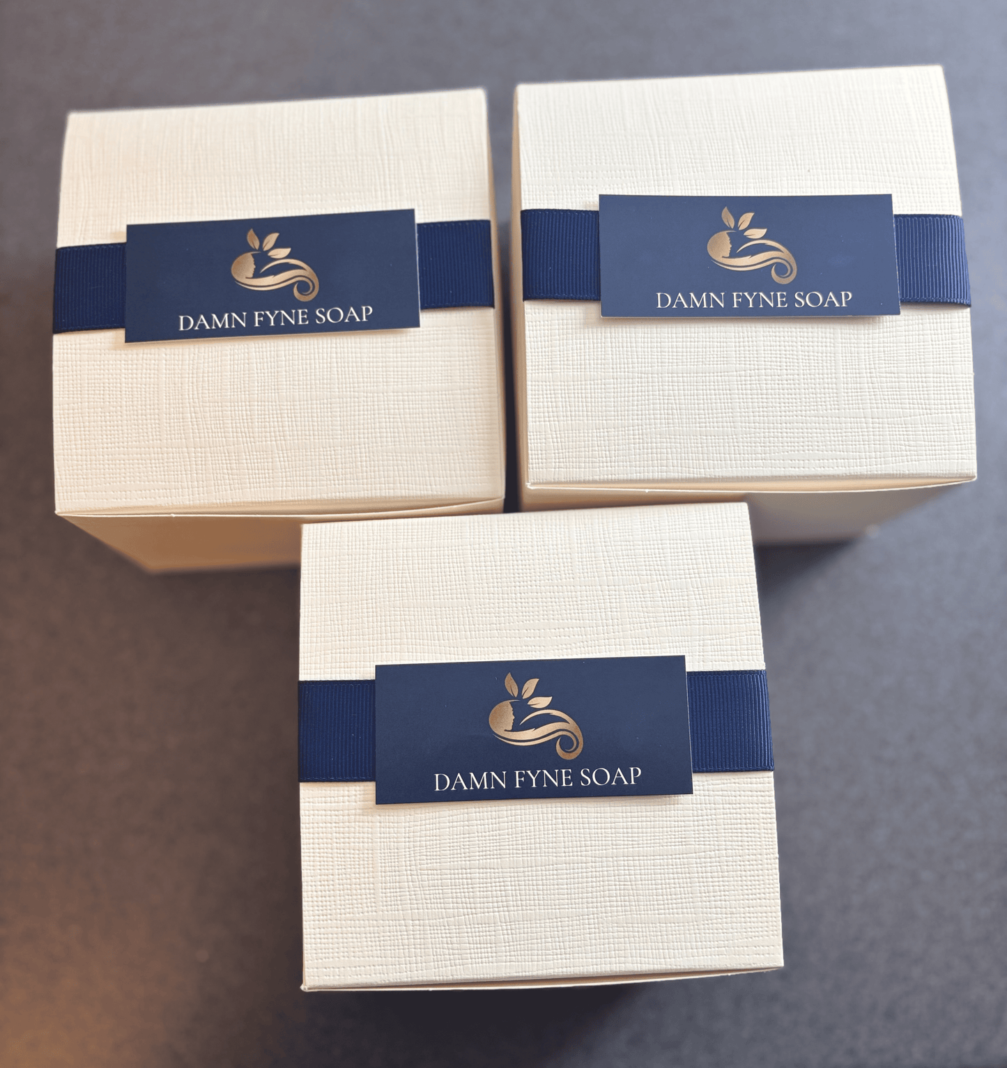 Cream Damn Fyne Soap gift box with navy ribbon, containing a guest-size handmade soap bar and a tin of whipped shea body butter. Styled with dried orange slice for festive presentation