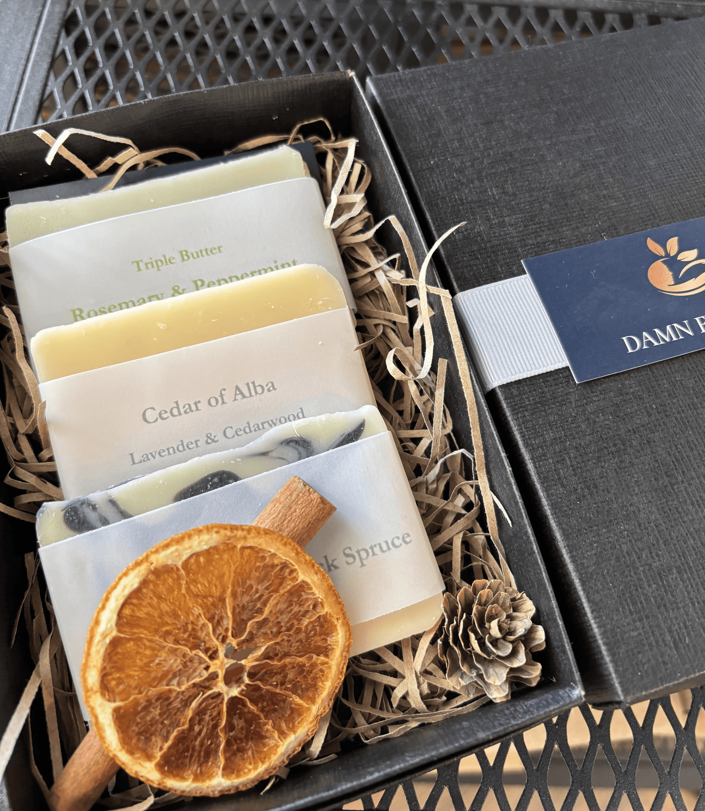 Elegant black gift box with festive styling, featuring three handmade soaps: Rosemary & Peppermint (triple butter), Cedar of Alba, and Cedarwood Fir & Black Spruce (triple butter). Nestled in shredded paper with dried orange slice, cinnamon stick, and pine cone for a rustic winter presentation