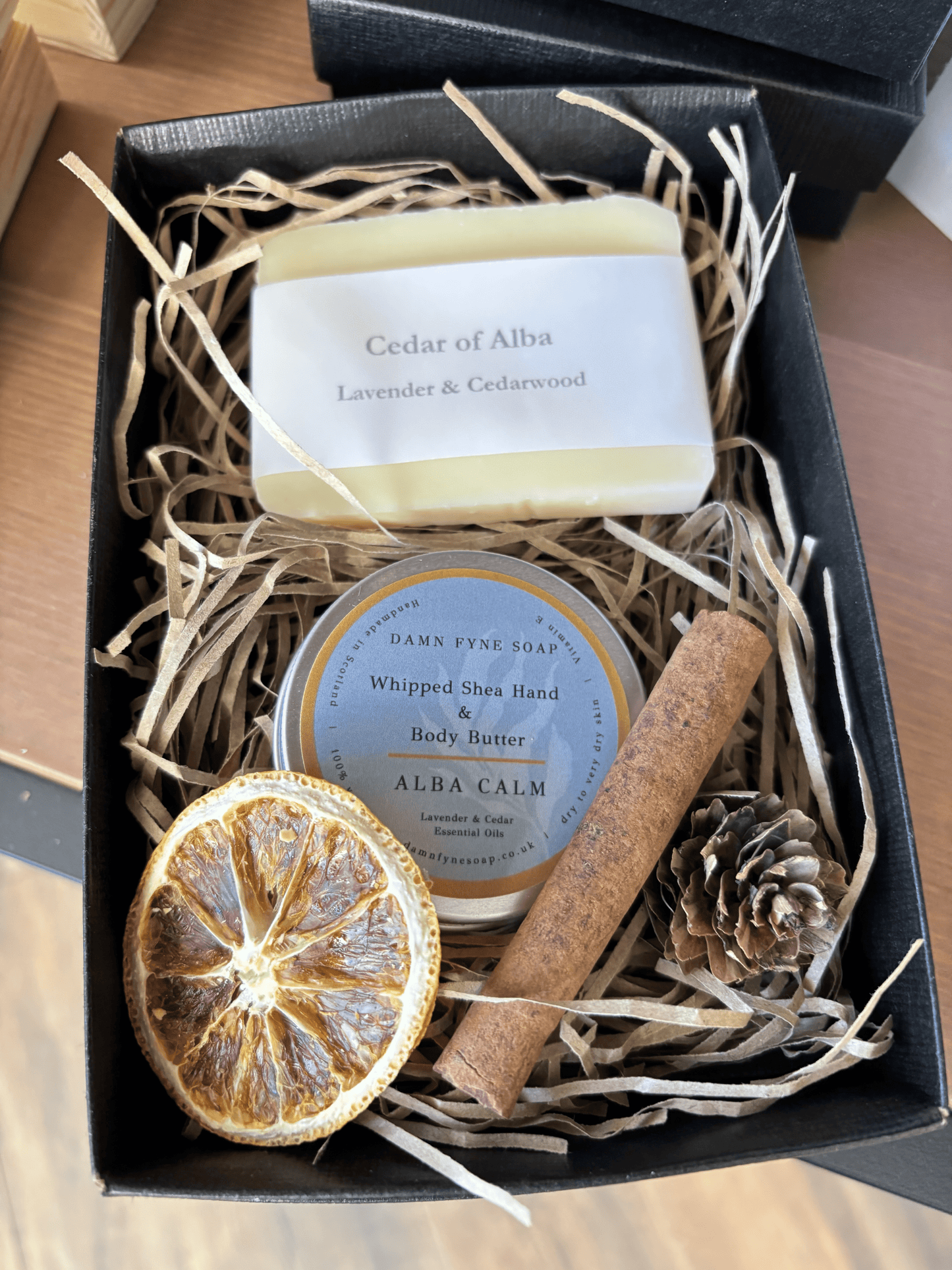 Black Damn Fyne Soap gift box with grey ribbon, containing Cedar of Alba soap and Alba Calm whipped shea body butter. Styled with dried orange slice, cinnamon stick, and pine cone for festive presentation