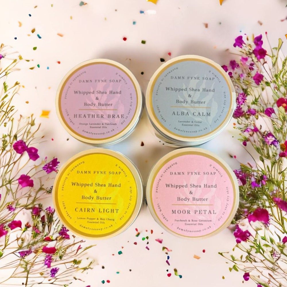 Four round containers of Damn Fyne Soap’s Whipped Shea Hand & Body Butters—Moor Petal, Heather Brae, Cairn Light, and Alba Calm—each with distinct coloured labels and essential oil blends. Handmade in Scotland for dry skin care.
