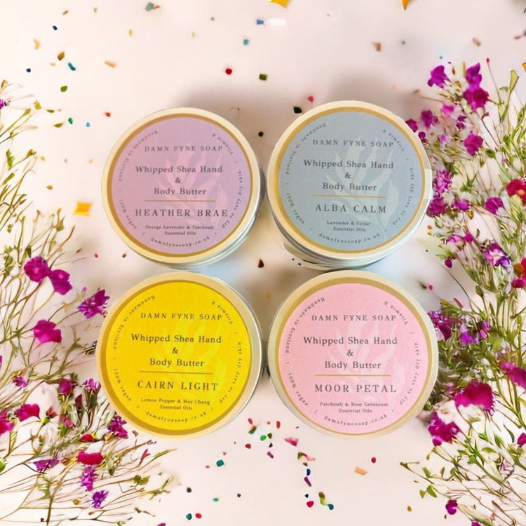 Four round containers of Damn Fyne Soap’s Whipped Shea Hand & Body Butters—Moor Petal, Heather Brae, Cairn Light, and Alba Calm—each with distinct coloured labels and essential oil blends. Handmade in Scotland for dry skin care.