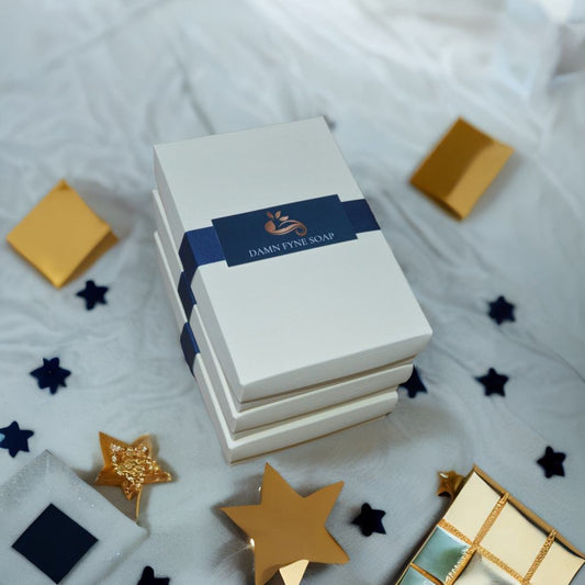 Elegant cream gift box from Damn Fyne Soap tied with dark blue ribbon, containing a trio of artisanal soaps in patchouli-based blends, styled with festive natural decorations.