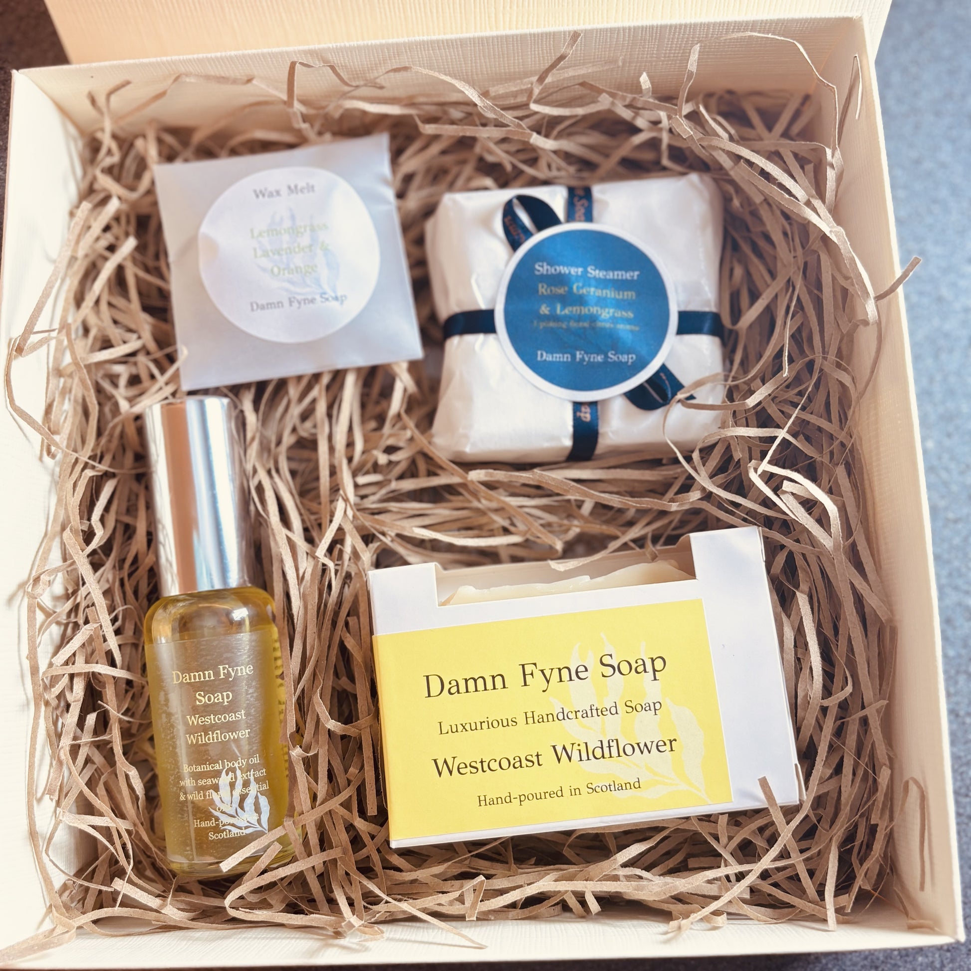 Cream gift box with navy ribbon containing Damn Fyne Soap products: Westcoast Wildflower soap, Westcoast Wildflower body oil, four shower steamers wrapped in white paper with dark ribbon, and a lemongrass, lavender & sweet orange wax melt, arranged on shredded paper filler for festive gifting
