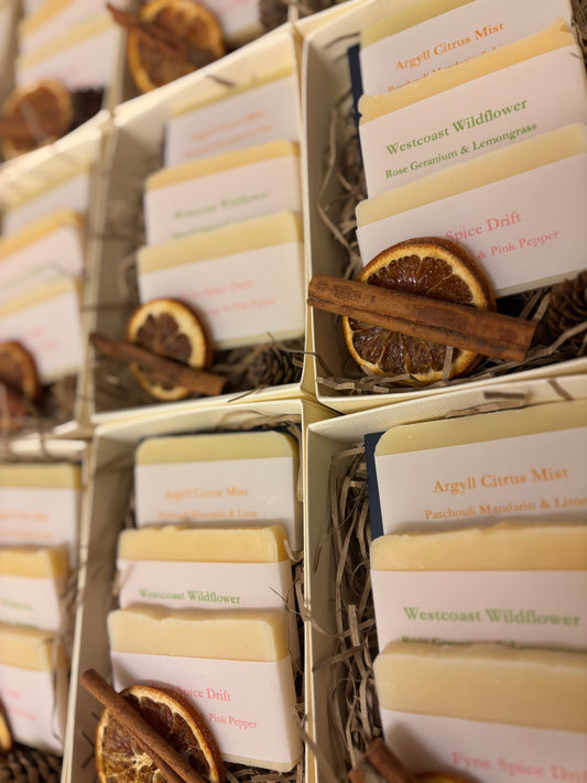 Festive Damn Fyne Soap gift box featuring three artisanal soap bars—Argyll Citrus Mist, Westcoast Wildflower, and Fyne Spice Drift—presented in a cream box with navy ribbon, dried orange slice, cinnamon stick, and pine cone.