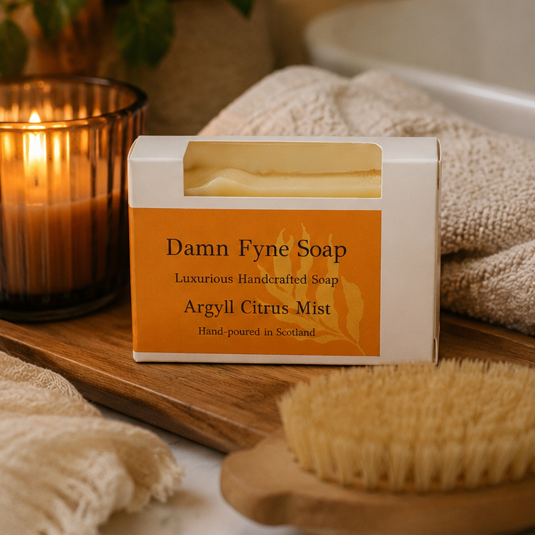 A boxed bar of soap labeled 'Damn Fyne Soap' and 'Argyll Citrus Mist' sits on a wooden tray with a lit candle, beige towels, and a brush in a warm, styled setting.