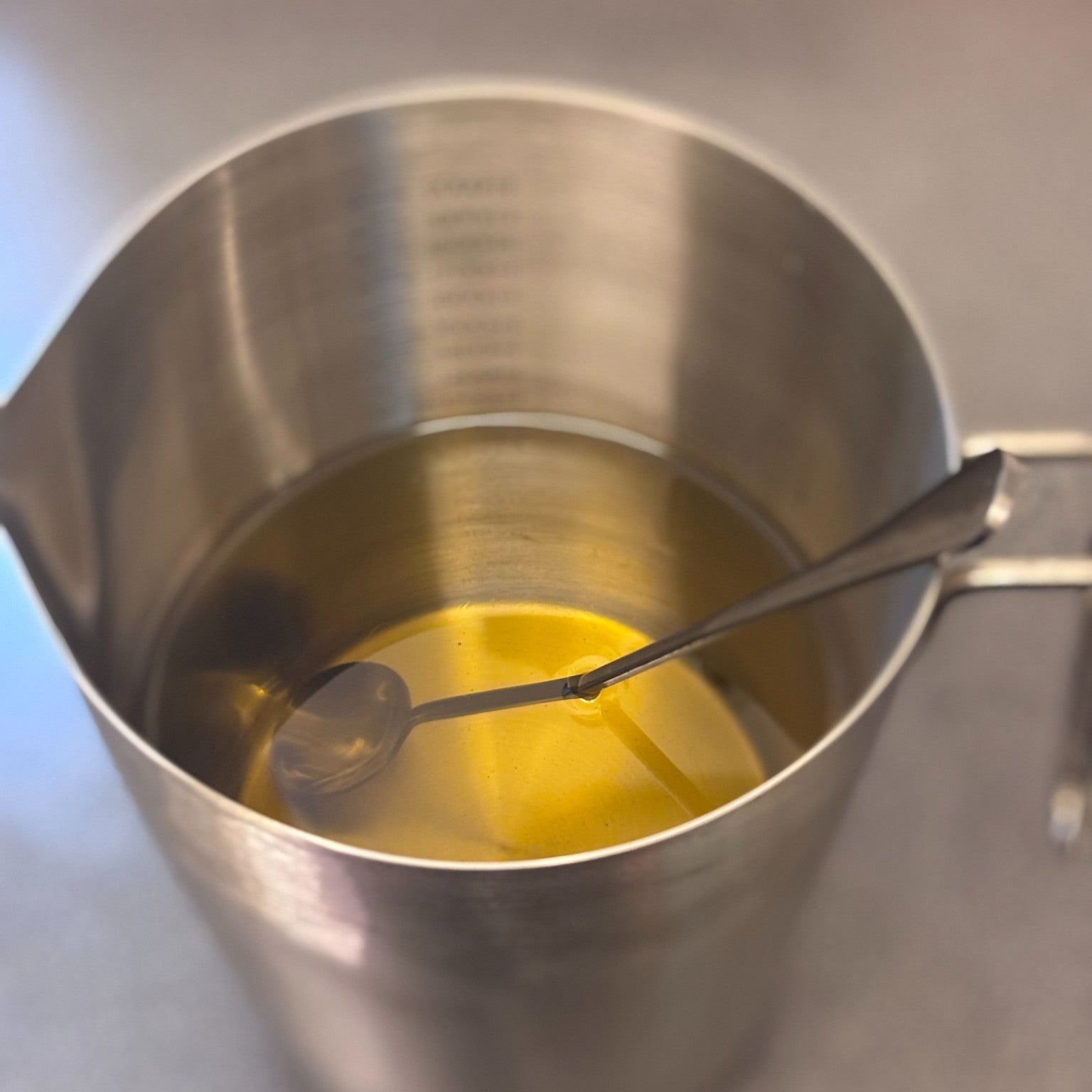 A stainless steel measuring pitcher filled with a pale yellow liquid, with a metal spoon resting inside, photographed from above on a neutral gray surface.