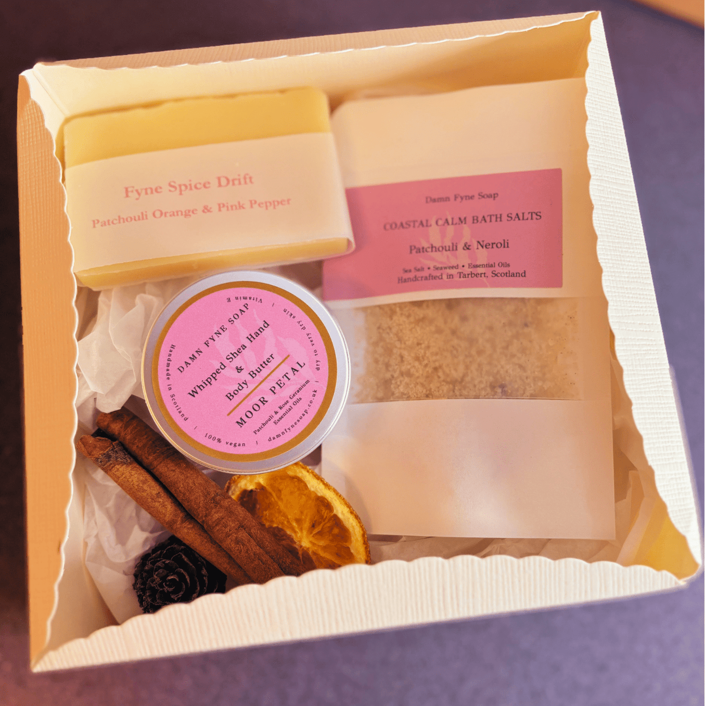 Cream gift box with navy ribbon, containing Fyne Spice Drift soap, Moor Petal body butter, and bath salts with patchouli & neroli. Styled with seasonal elements like dried orange slice 