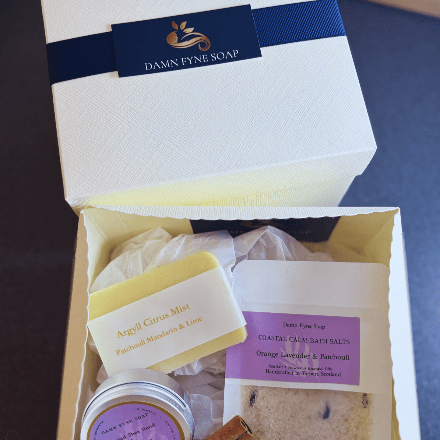 Cream Damn Fyne Soap gift box with navy ribbon, containing guest-size soap, whipped body butter, and bath salts sachet. Styled with dried orange slice for festive presentation 