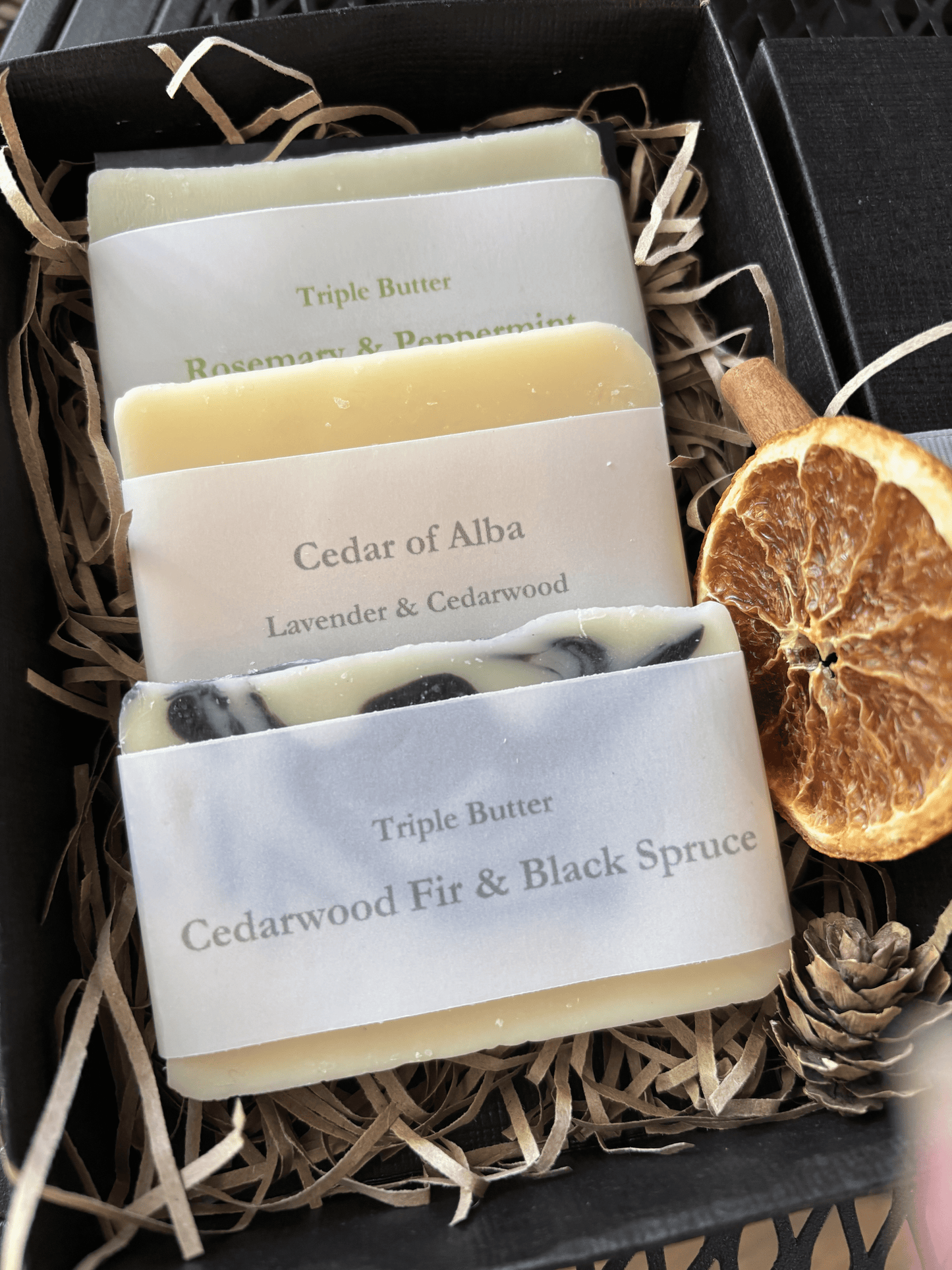 Black Damn Fyne Soap gift box with festive styling, containing three handmade soap bars: Rosemary & Peppermint (triple butter), Cedar of Alba, and Cedarwood Fir & Black Spruce (triple butter). Styled with dried orange slice, cinnamon stick, and pine cone 