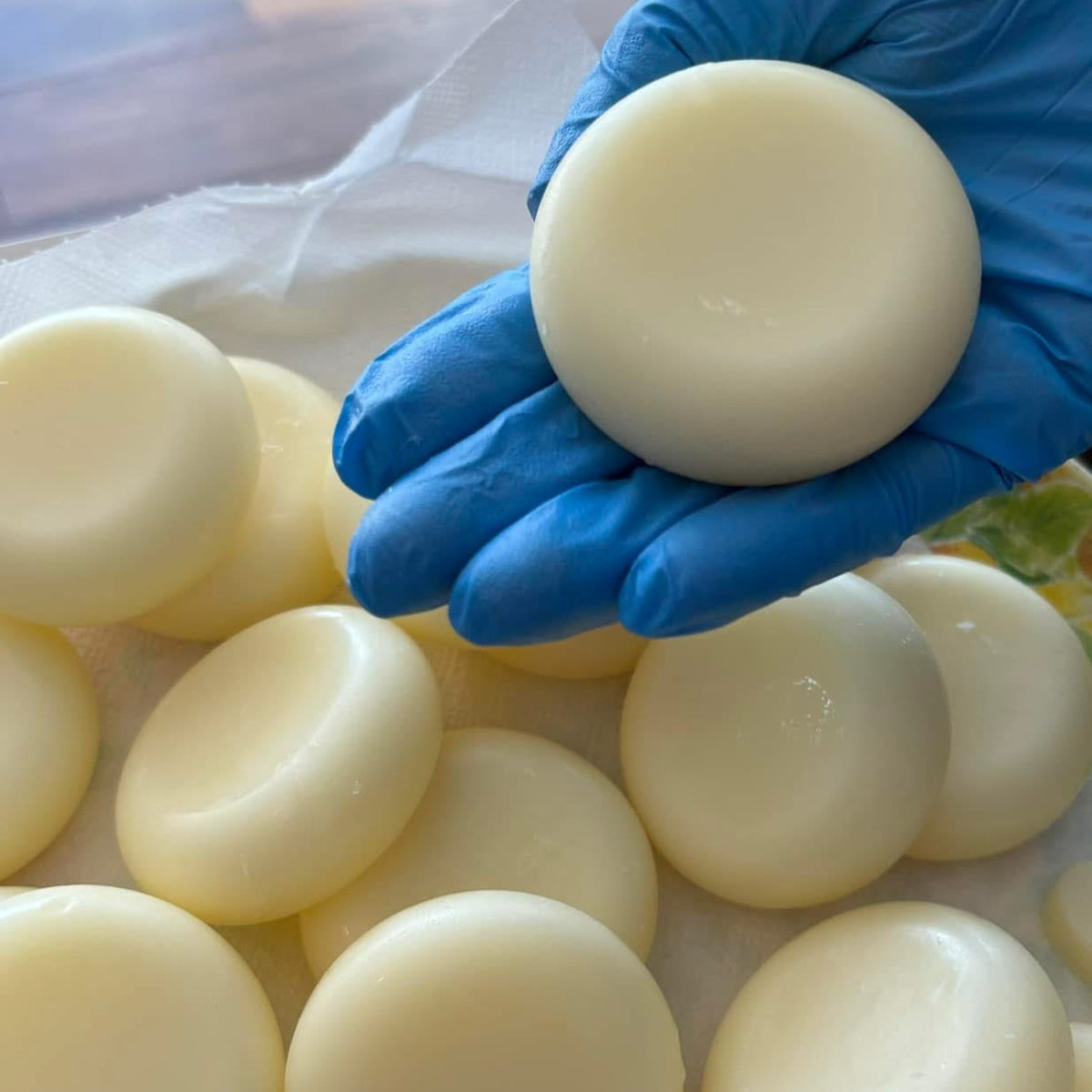 A hand wearing a blue nitrile glove holds a smooth, round, off-white bar of solid conditioner bar above a pile of similar bars on a white cloth.