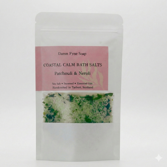 Coastal Calm Bath Salts in a semi-transparent kraft pouch with a soft pink label. Label reads “Coastal Calm Bath Salts – Patchouli & Neroli – Sea Salt + Seaweed + Essential Oils – Handcrafted in Tarbert, Scotland.” Contents visible through window, showing coarse green and white bath salts with flecks of seaweed