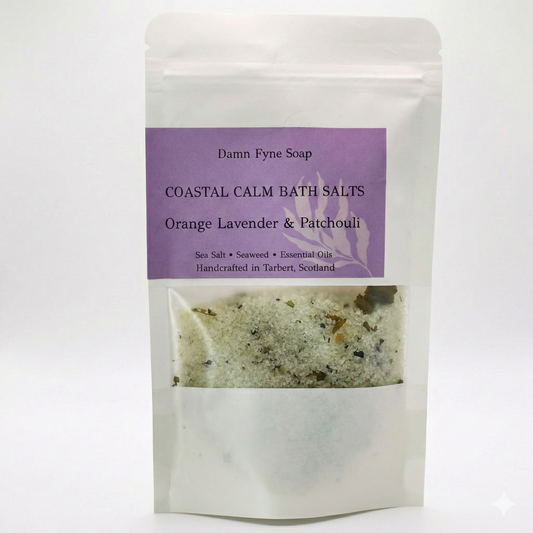 Coastal Calm Bath Salts in a semi-transparent kraft pouch with a purple label. Label reads “Coastal Calm Bath Salts – Orange, Lavender & Patchouli – Sea Salt • Seaweed • Essential Oils – Handcrafted in Tarbert, Scotland.” Contents visible through window, showing coarse salt and botanical elements.